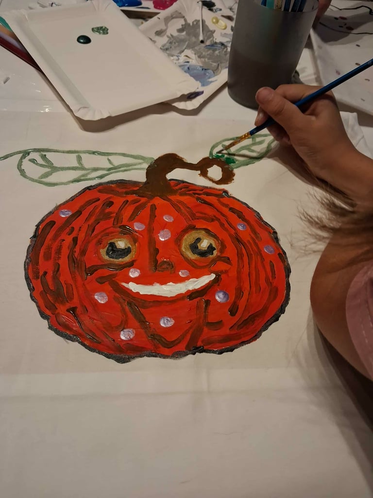 Child painting a smiling red pumpkin with a brush at a white craft table with supplies
